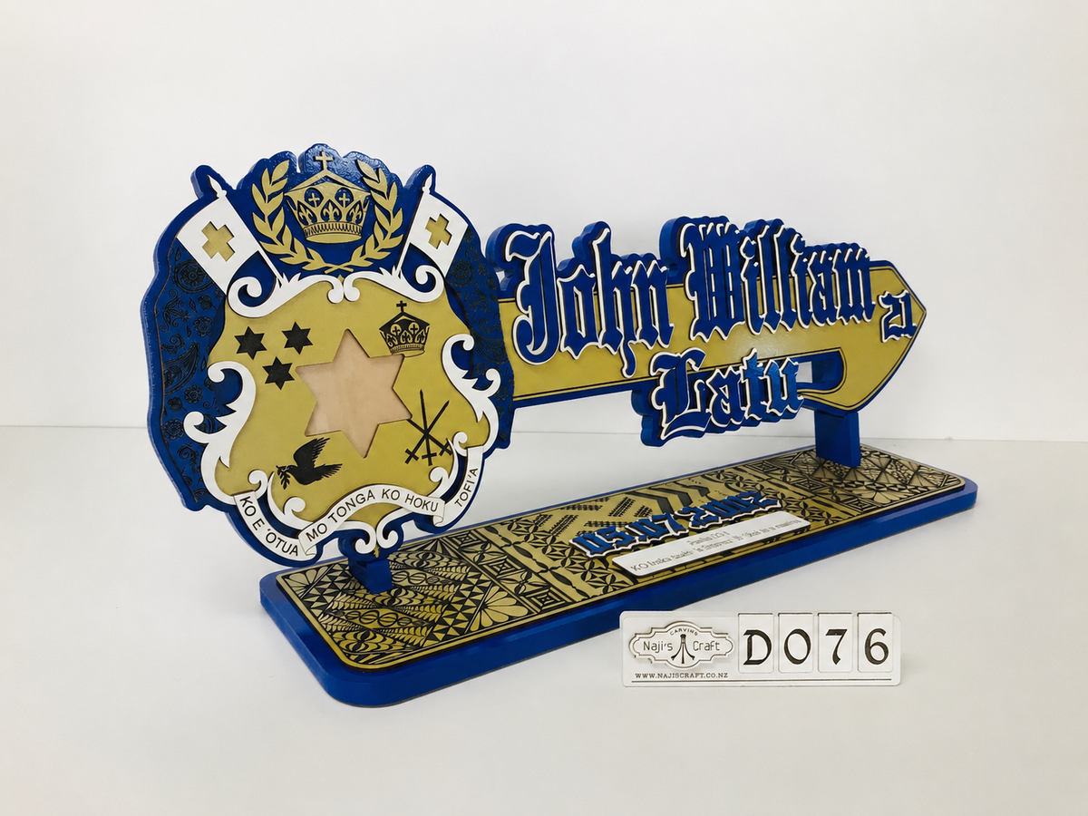 A decorative Tongan 21st birthday wooden key or display stand featuring a blue and gold Tongan emblem with the name 'John William Bette' in stylized lettering, mounted on a long base with ornate gold detailing. Key theme number is D076.